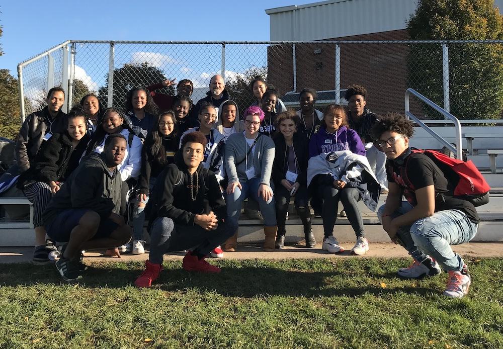 A diverse group of students and faculty members posing together on outdoor bleachers, with trees and a school building in the background, conveying a sense of community and school spirit.